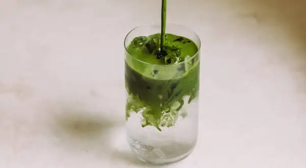 Ein leuchtend grüner Matcha-Sirup fließt in ein Glas mit Eis und sprudelndem Wasser. Ein erfrischender Matcha Tonic entsteht.