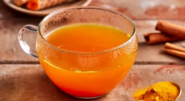 Ein Glas goldener Kurkuma Tee, daneben frische Kurkumawurzeln, Zimtstangen und ein Löffel Kurkumapulver. So einfach lässt sich Kurkuma Tee zubereiten.