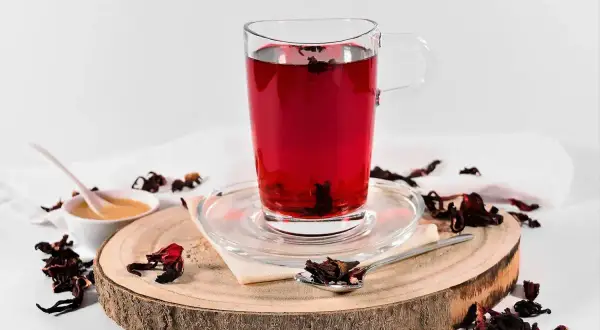 Ein Glas tiefroter Hibiskus-Tee, bereit zur Zubereitung. Getrocknete Hibiskusblüten und Honig sind daneben arrangiert.
