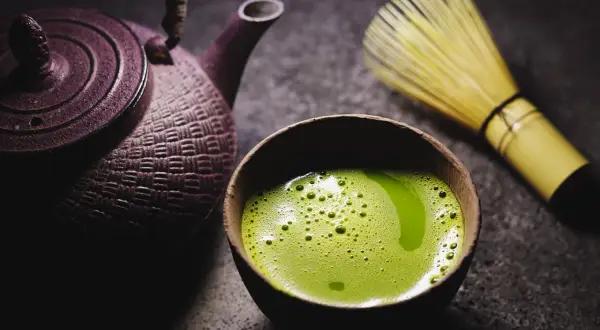 Eine traditionelle japanische Teekanne und ein Chasen (Bambusbesen) neben einer Schale mit schaumigem, leuchtend grünem Matcha. So wird Matcha zubereitet.