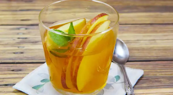 Ein Glas mit goldenem kamillentee mit honig, garniert mit Apfelscheiben und Minze, steht auf einem Holztisch.