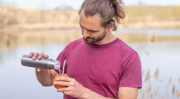 Mann mit Dutt und Bart gießt heißes Wasser aus Thermoskanne in Kalebasse für Mate oder Matcha.