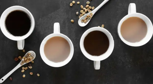 Vier Tassen Kaffee, zwei schwarz, zwei mit Milch, und Löffel mit Kaffeebohnen. Die Entscheidung zwischen Tee vs Kaffee ist hier klar: Kaffee!
