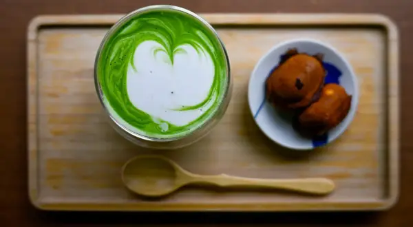 Ein Glas cremiger Matcha mit Herz-Latté-Art und zwei süßen Leckereien. Perfekt zum Matcha trinken.