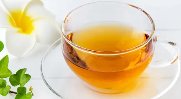 Klarer schwarzer Tee in einer Glas Tasse, beruhigend für den Magen, neben einer weißen Blüte und grünen Blättern.