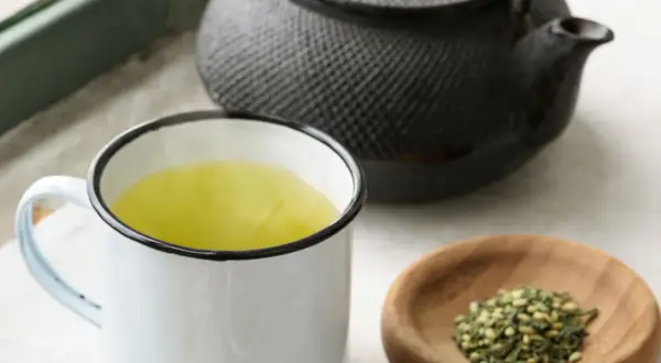 Dampfender Genmaicha in einer Tasse, daneben eine Teekanne und eine Schale mit geröstetem Reis. Die Genmaicha Zubereitung ist im Gange.