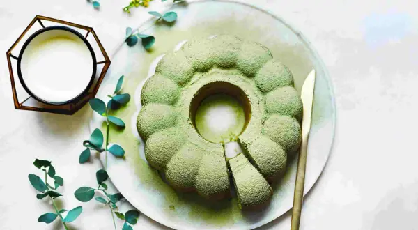 Ein köstlicher veganer Matcha-Kuchen, bestäubt mit Puderzucker, liegt auf einem Teller. Daneben ein Glas mit weißer Flüssigkeit und grünes Blattwerk.