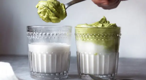 Ein Löffel mit cremigem Dalgona Matcha wird über ein Glas Milch gehalten. Daneben steht ein fertiges Glas Dalgona Matcha.