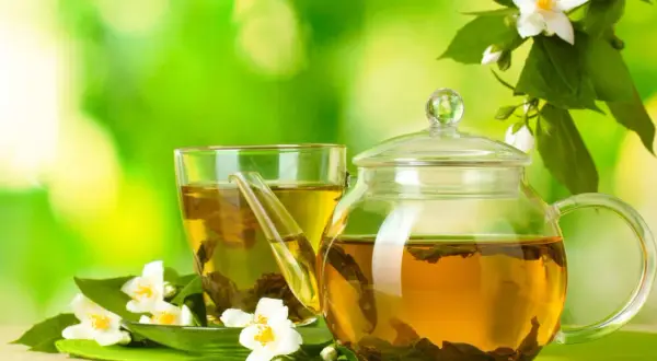 Glas-Teekanne und Tasse mit goldenem Jasmin Tee, umgeben von frischen Jasminblüten. Perfekt für die Jasmin Tee Zubereitung.