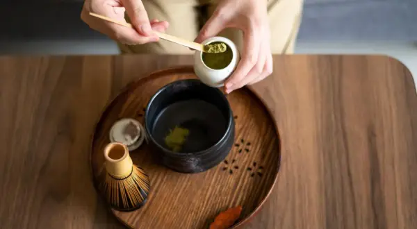 Vorbereitung von Matcha für die Schwangerschaft: Hände schöpfen grünes Pulver in eine Schale mit heißem Wasser. Ein Chasen (Bambusbesen) liegt daneben.