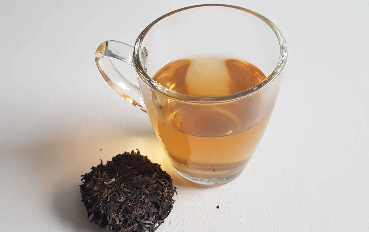 Ein Glas goldbrauner Pu-erh-Tee neben trockenen Teeblättern. Der Tee liefert sanftes Koffein für einen belebenden Start.