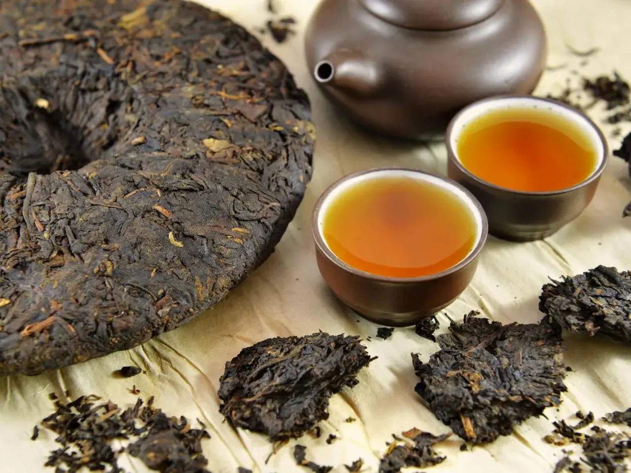 Ein runder Kuchen und lose Blätter von dunklem Pu-Erh-Tee neben zwei Tassen des bernsteinfarbenen Getränks und einer Teekanne.