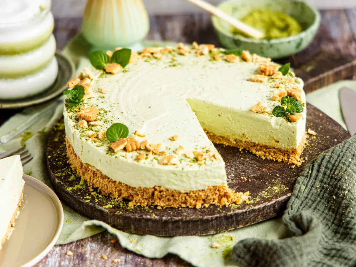 Ein cremiger Matcha Cheesecake mit Keksboden, garniert mit Minze und zerbröselten Keksen. Ein Stück wurde bereits angeschnitten.