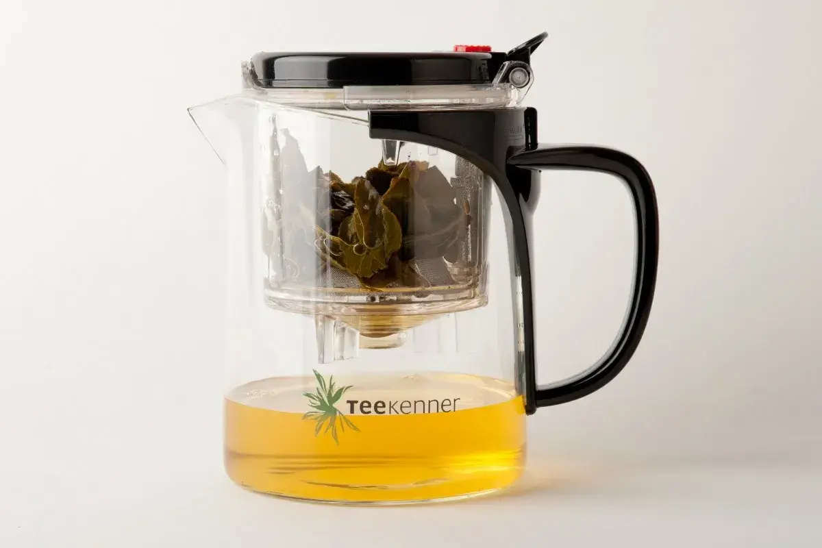 Glas-Teekanne mit schwarzem Deckel und Griff, gefüllt mit goldenem Tee und grünen Teeblättern. Perfekt, um Tee zu servieren.
