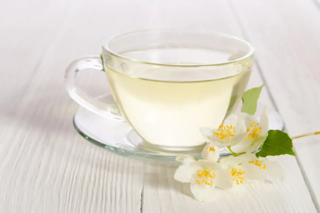 Eine Tasse klarer, heller Flüssigkeit, die an weißen Tee erinnert, mit duftenden Jasminblüten daneben.