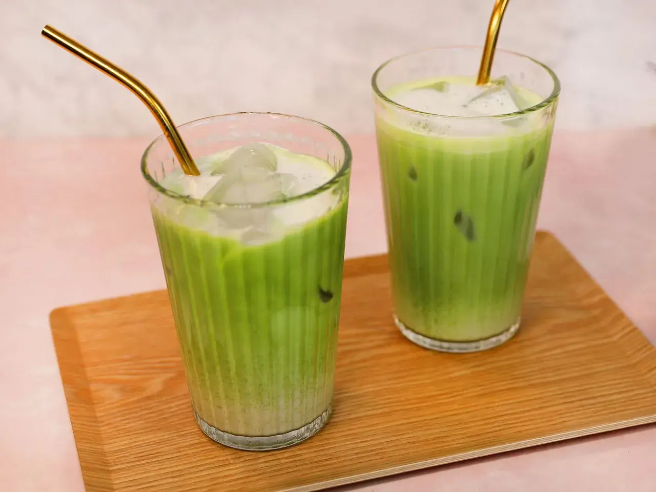 Zwei Gläser mit erfrischendem Matcha Hafermilch-Eisgetränk, serviert auf einem Holztablett. Goldene Strohhalme ragen aus den Getränken.