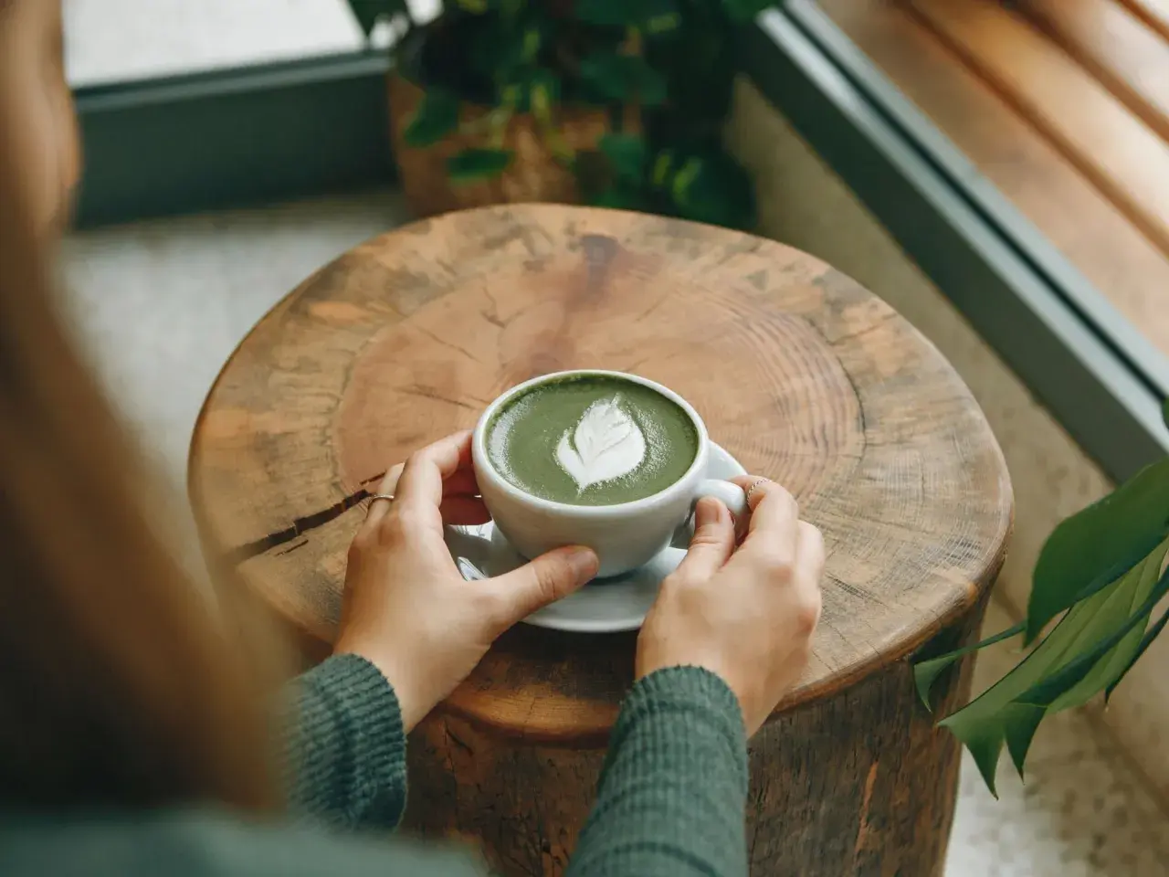 Hände halten eine Tasse cremigen Matcha Latte mit Latte Art auf einem rustikalen Holztisch. Eine gesunde Alternative zu Kaffee.