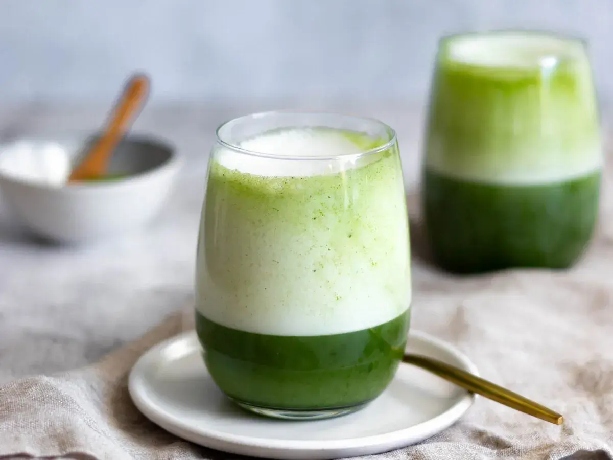 Zwei Gläser mit geschichtetem Matcha-Latte, die die perfekte Matcha-Zubereitung zeigen. Ein Löffel liegt daneben.