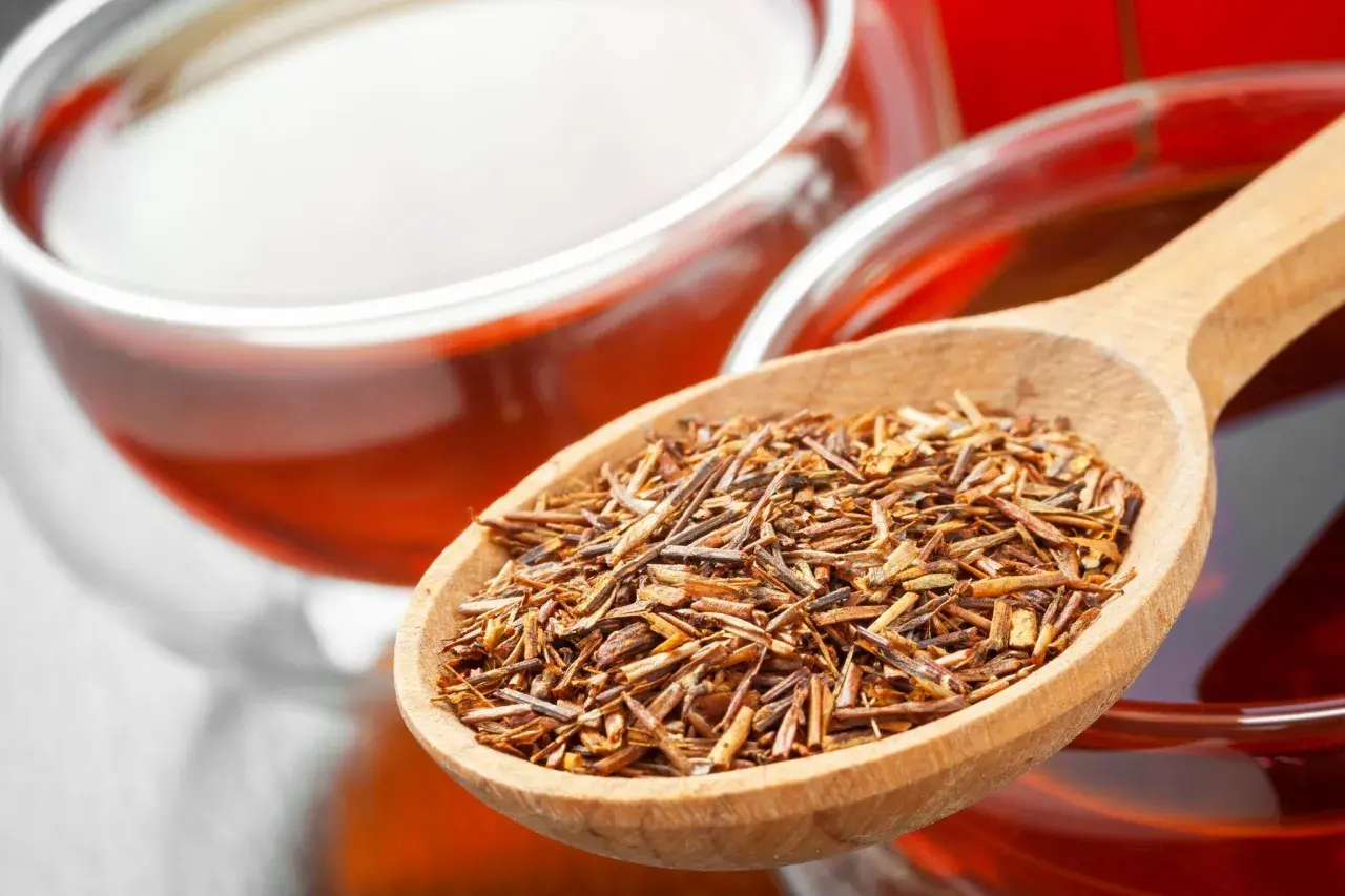 Rooibos Tee: Wirkung Schlafen. Eine Holzschaufel voller Rooibos-Blätter neben einer Tasse des roten Tees.