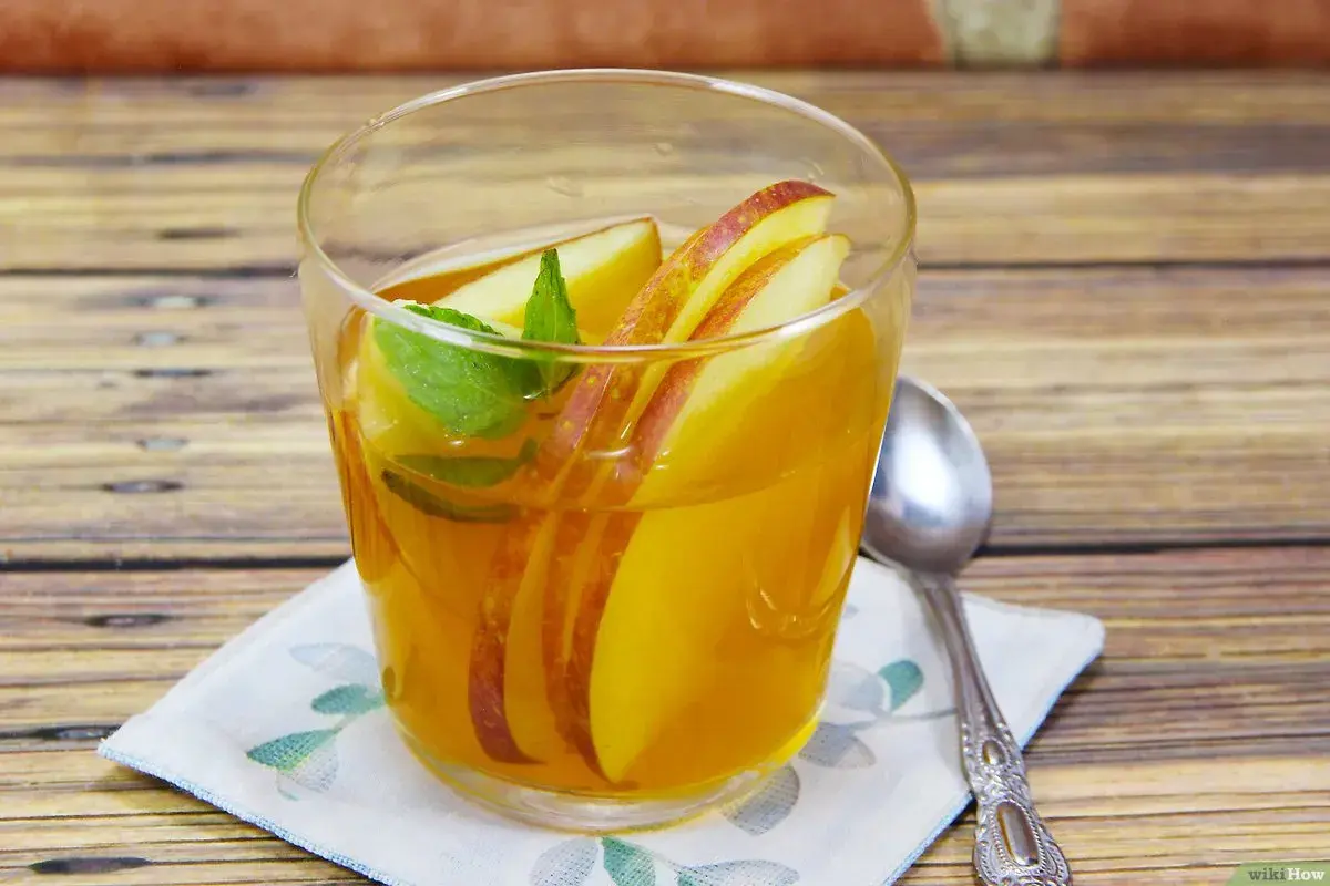 Ein Glas mit goldenem kamillentee mit honig, garniert mit Apfelscheiben und Minze, steht auf einem Holztisch.