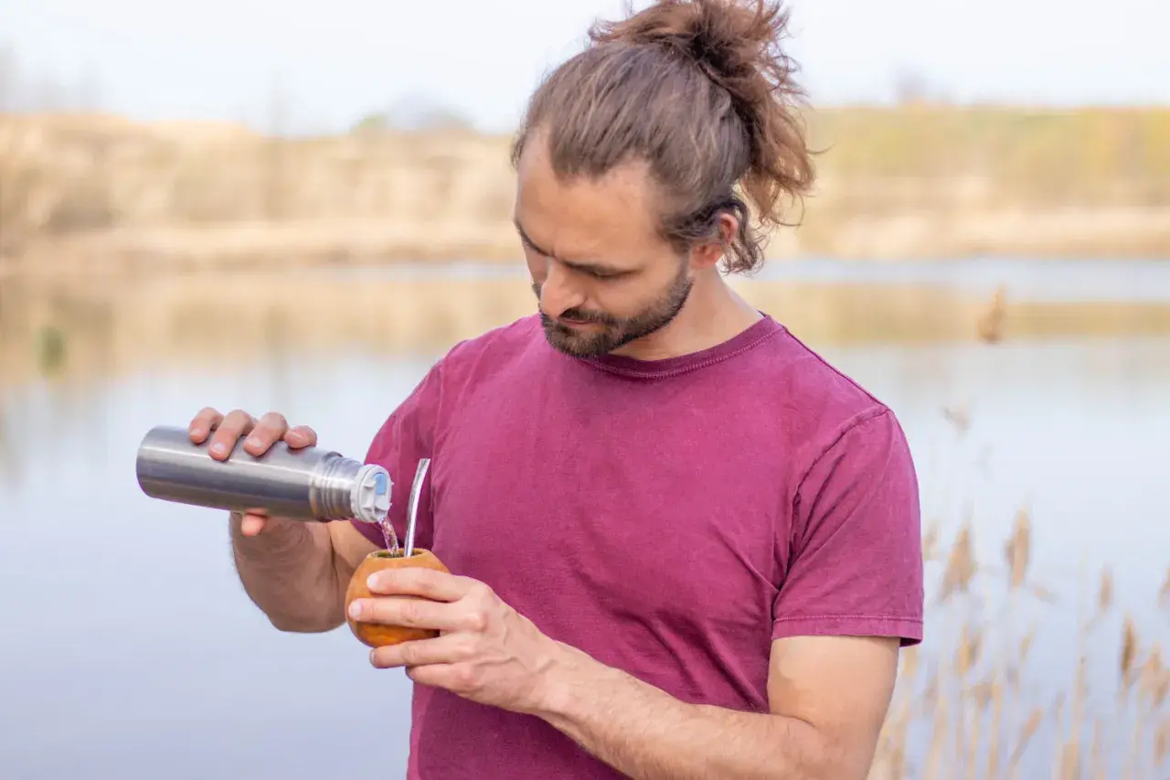 Mann mit Dutt und Bart gießt heißes Wasser aus Thermoskanne in Kalebasse für Mate oder Matcha.