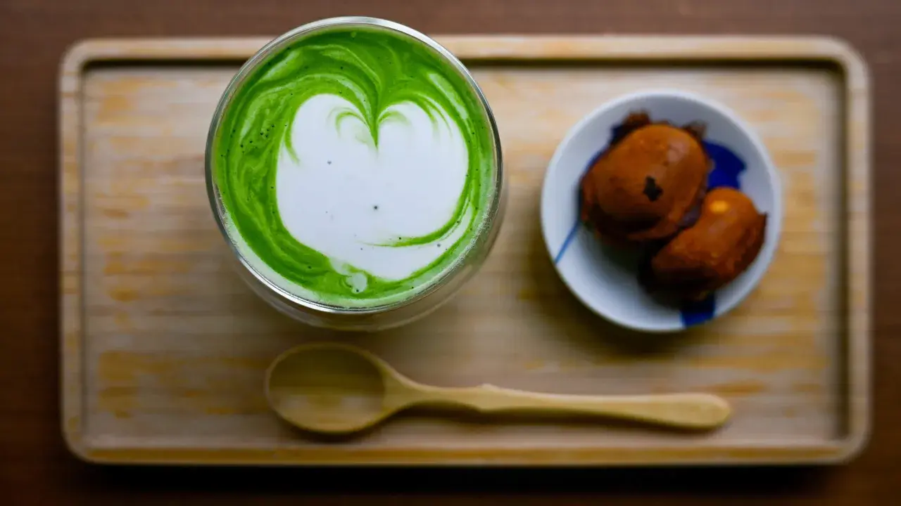 Ein Glas cremiger Matcha mit Herz-Latté-Art und zwei süßen Leckereien. Perfekt zum Matcha trinken.