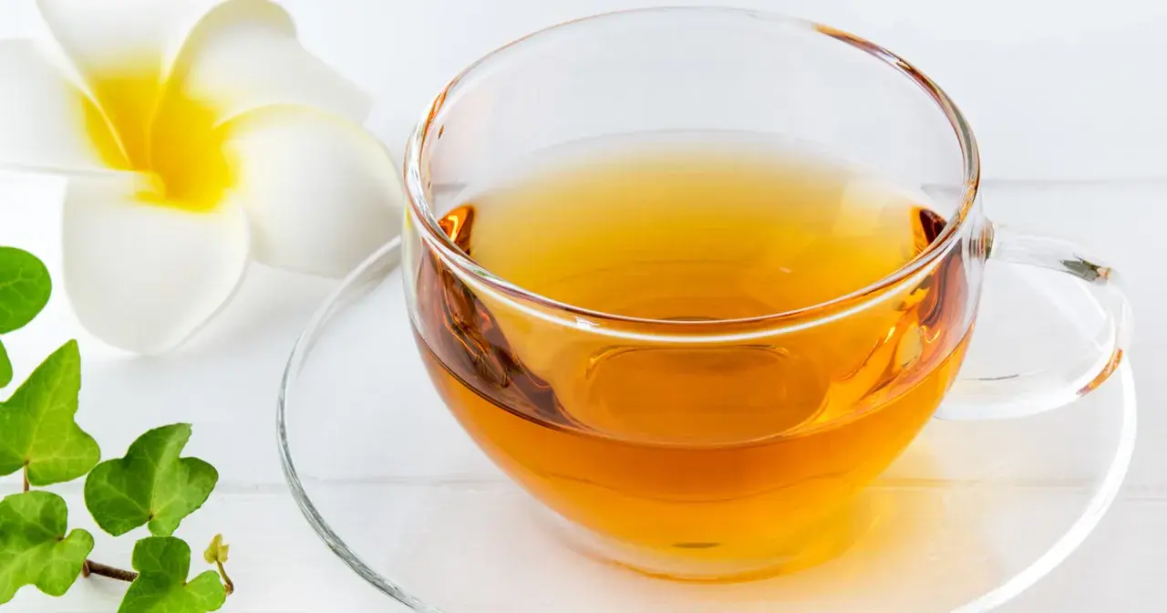 Klarer schwarzer Tee in einer Glas Tasse, beruhigend für den Magen, neben einer weißen Blüte und grünen Blättern.