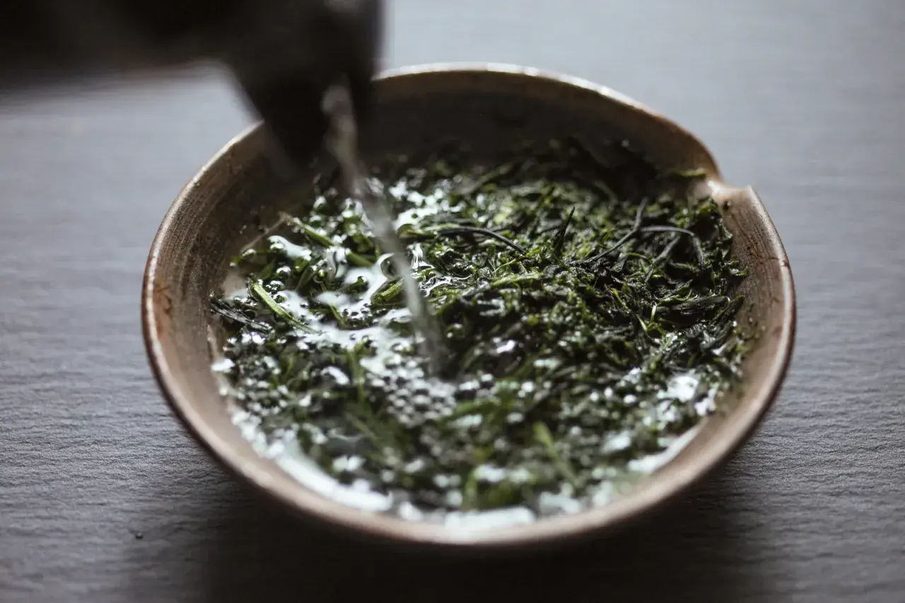 Wasser fließt in eine Schale mit grünen Teeblättern für die Gyokuro Zubereitung.