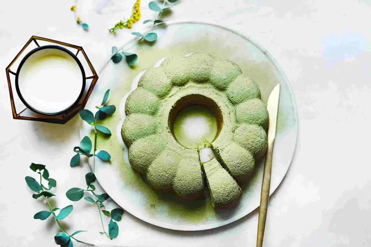 Ein köstlicher veganer Matcha-Kuchen, bestäubt mit Puderzucker, liegt auf einem Teller. Daneben ein Glas mit weißer Flüssigkeit und grünes Blattwerk.
