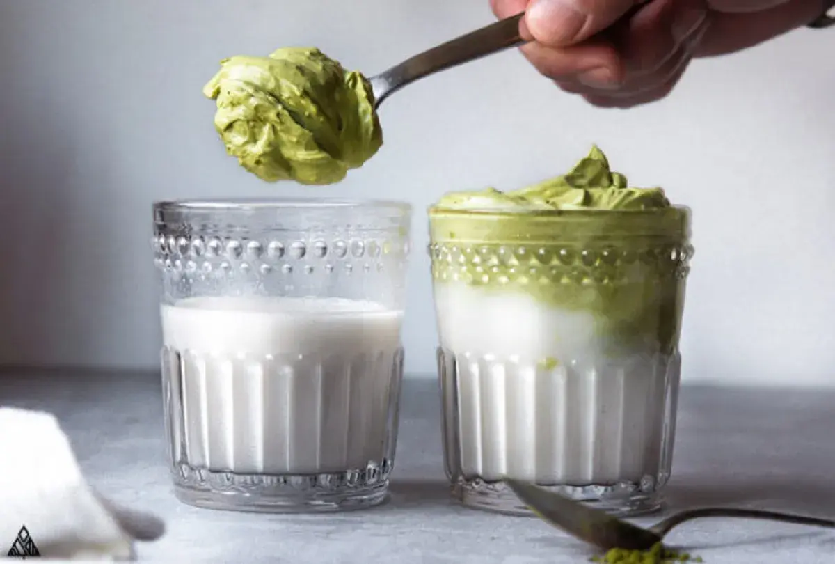 Ein Löffel mit cremigem Dalgona Matcha wird über ein Glas Milch gehalten. Daneben steht ein fertiges Glas Dalgona Matcha.