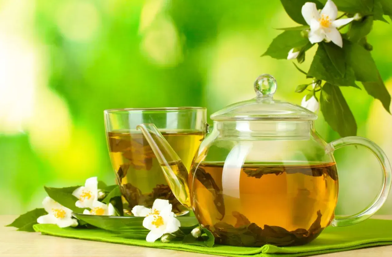 Glas-Teekanne und Tasse mit goldenem Jasmin Tee, umgeben von frischen Jasminblüten. Perfekt für die Jasmin Tee Zubereitung.