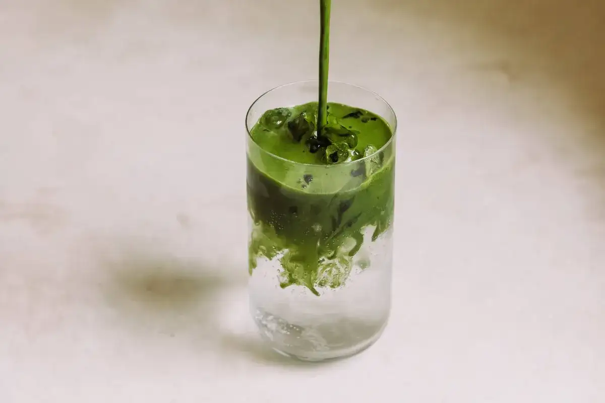 Ein leuchtend grüner Matcha-Sirup fließt in ein Glas mit Eis und sprudelndem Wasser. Ein erfrischender Matcha Tonic entsteht.