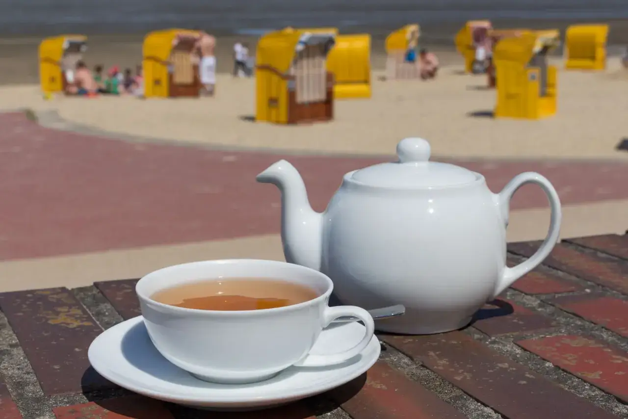 Ein wei&szlig;es Teeservice mit einer Tasse Tee steht auf einem Tisch. Im Hintergrund sind gelbe Strandk&ouml;rbe und Menschen am Strand zu sehen. Perfekt, um zu &uuml;berlegen, welchen Tee man morgens trinken m&ouml;chte.