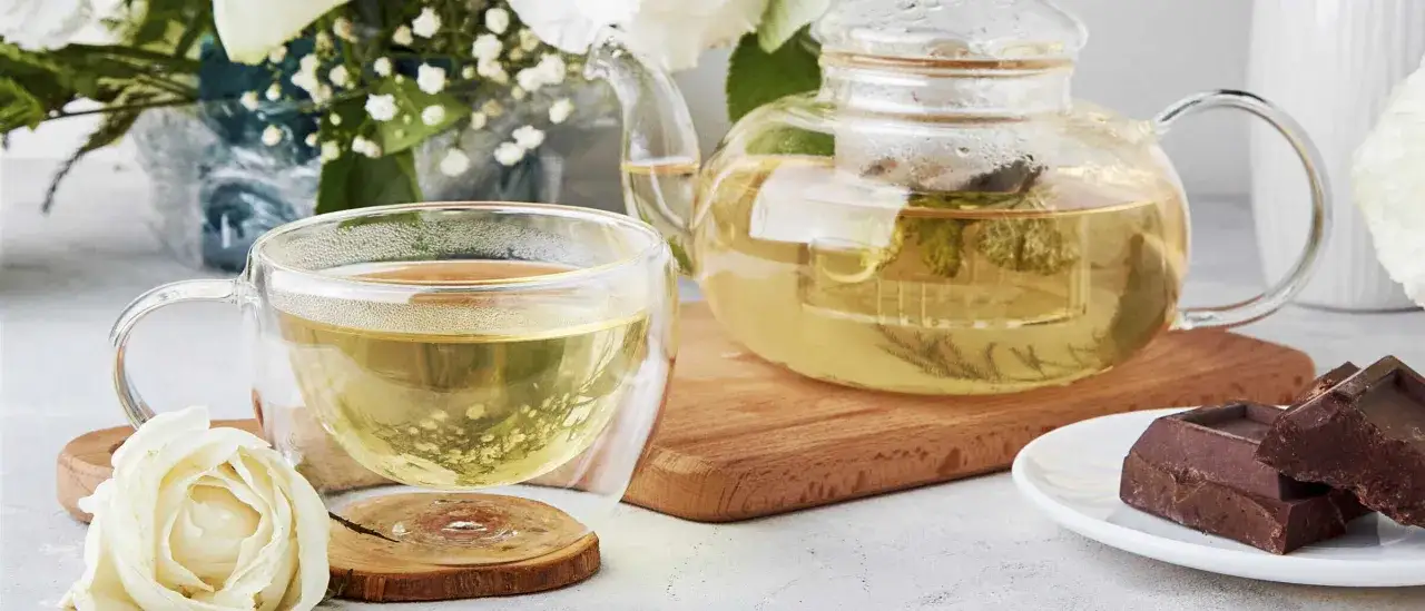 Ein Glas und eine Kanne mit hellem, duftendem Tee, daneben Schokolade und eine wei&szlig;e Rose. So sieht Genuss aus, was ist weisser tee?