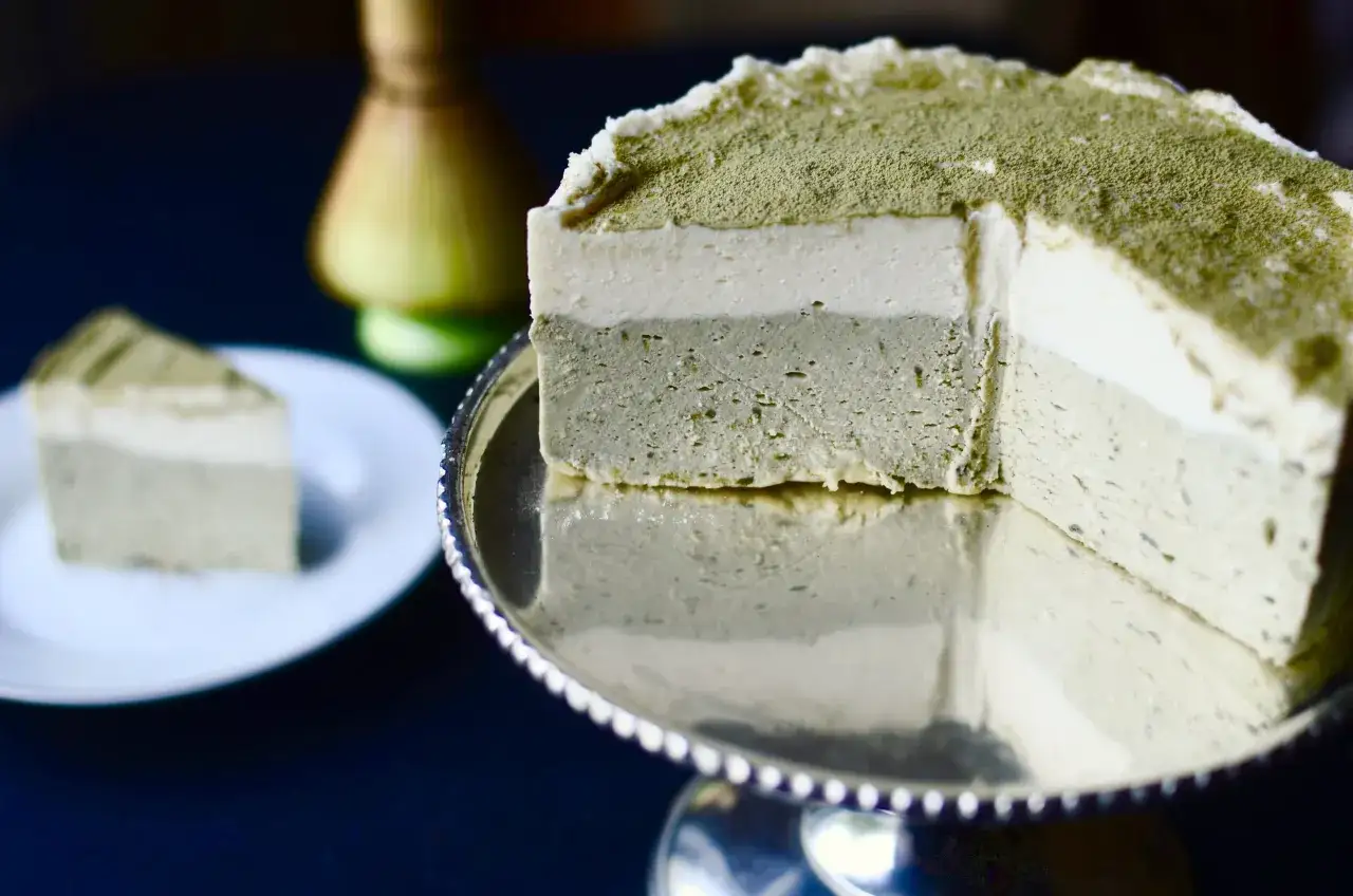 Schichtiger Matcha Cheesecake auf einer silbernen Tortenplatte, best&auml;ubt mit gr&uuml;nem Pulver. Ein St&uuml;ck liegt auf einem wei&szlig;en Teller.