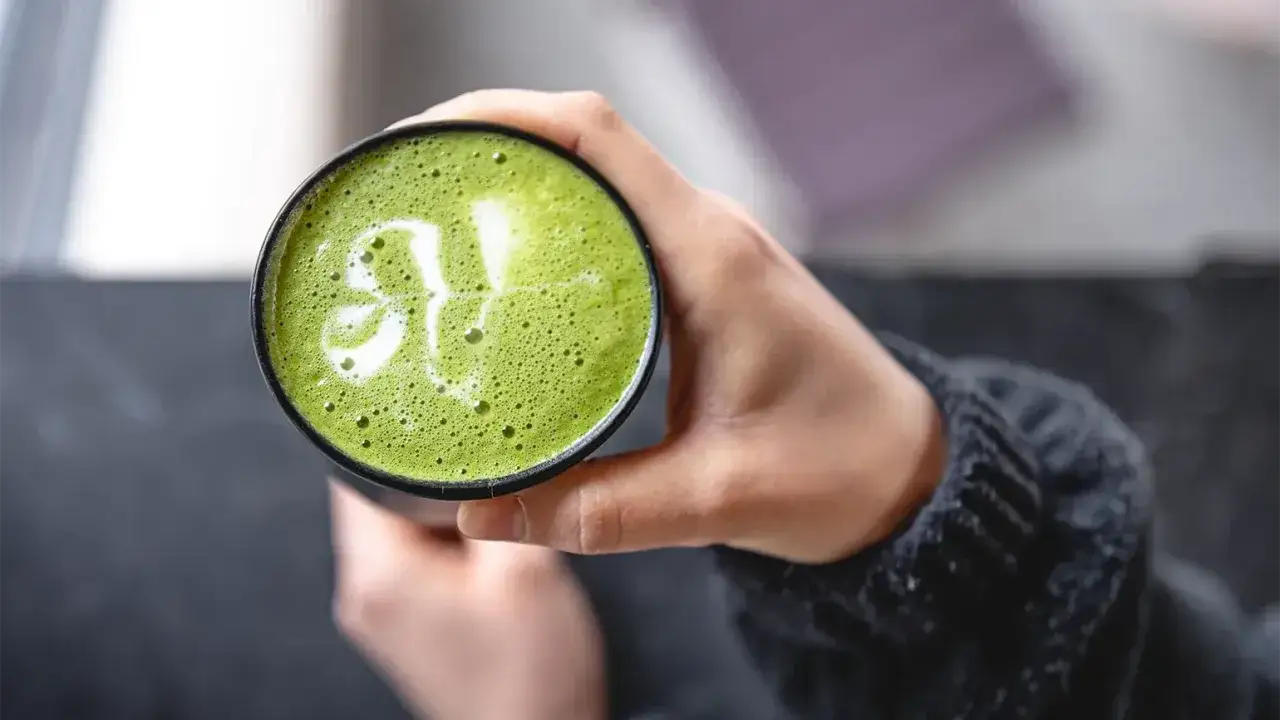 H&auml;nde halten eine Tasse mit gr&uuml;nem Matcha-Latte, verziert mit Milchschaum. F&uuml;r manche ist Matcha histaminarm, eine Wohltat.