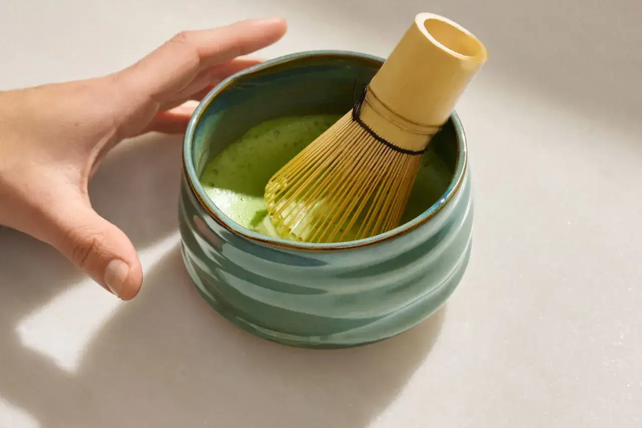 Hand bereitet cremigen Matcha mit einem Chasen in einer grünen Schale zu.