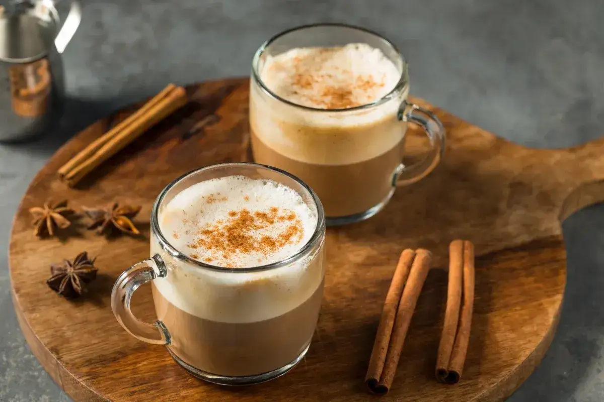 Zwei Tassen cremiger Rooibos Tee mit Milch, bestreut mit Zimt, auf einem Holzbrett mit Zimtstangen und Sternanis.