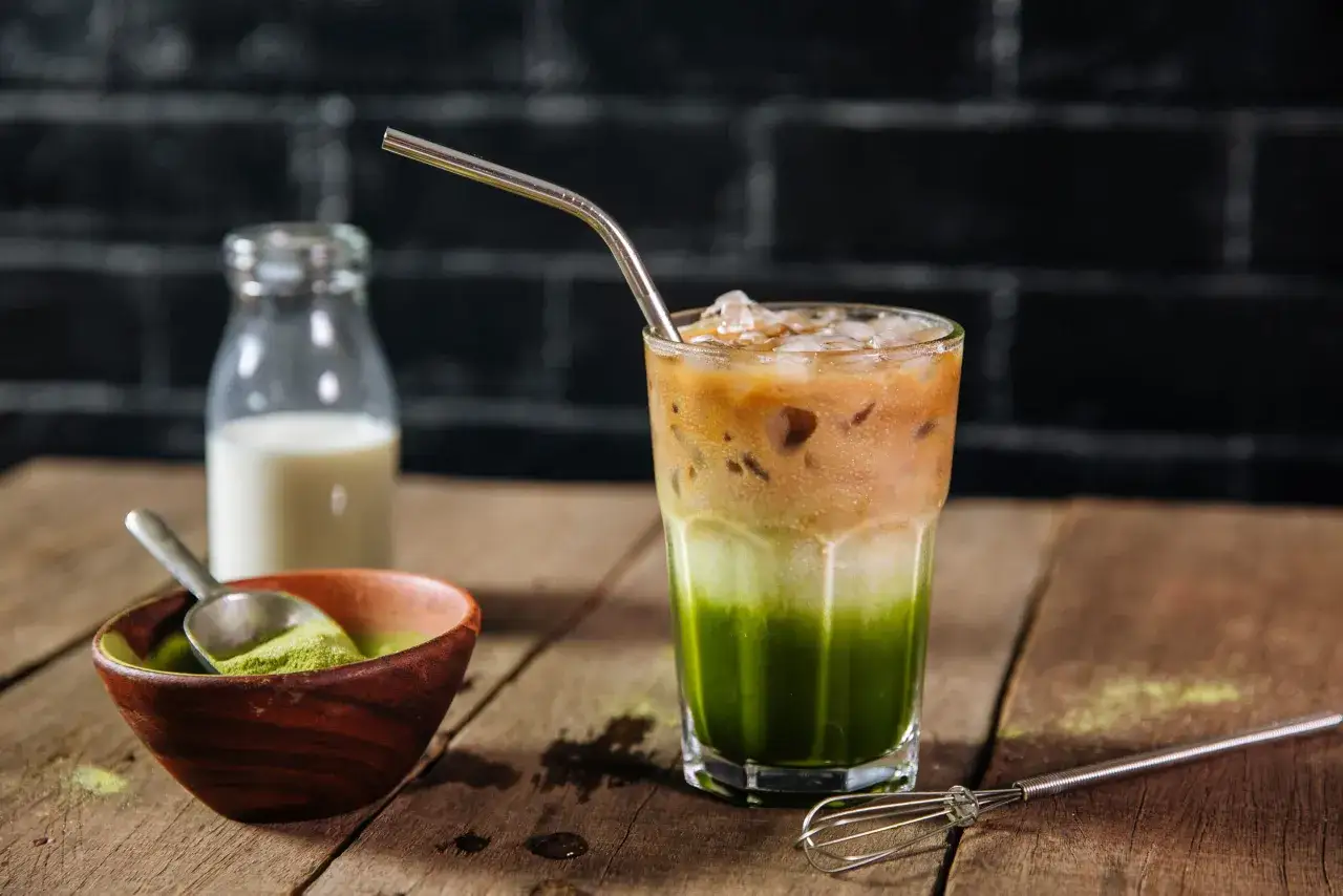 Ein geschichteter Eiskaffee mit Matcha, Milch und Eis. Daneben eine Schale mit Matcha-Pulver und eine Milchflasche.