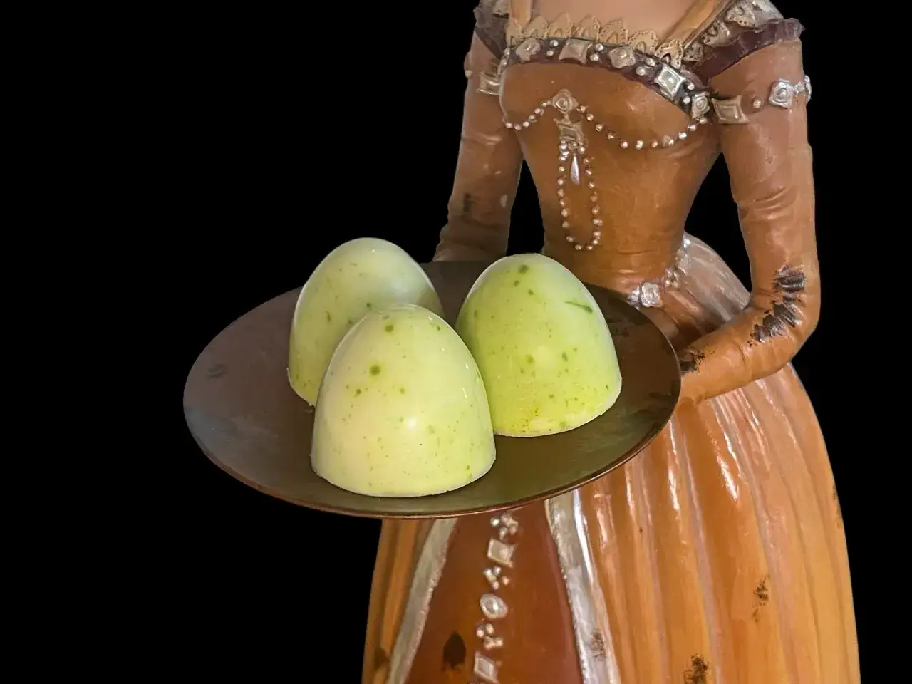 Drei gr&uuml;ne matcha pralinen mit Tupfen auf einer braunen Platte, gehalten von einer Figur in einem orangefarbenen Kleid.