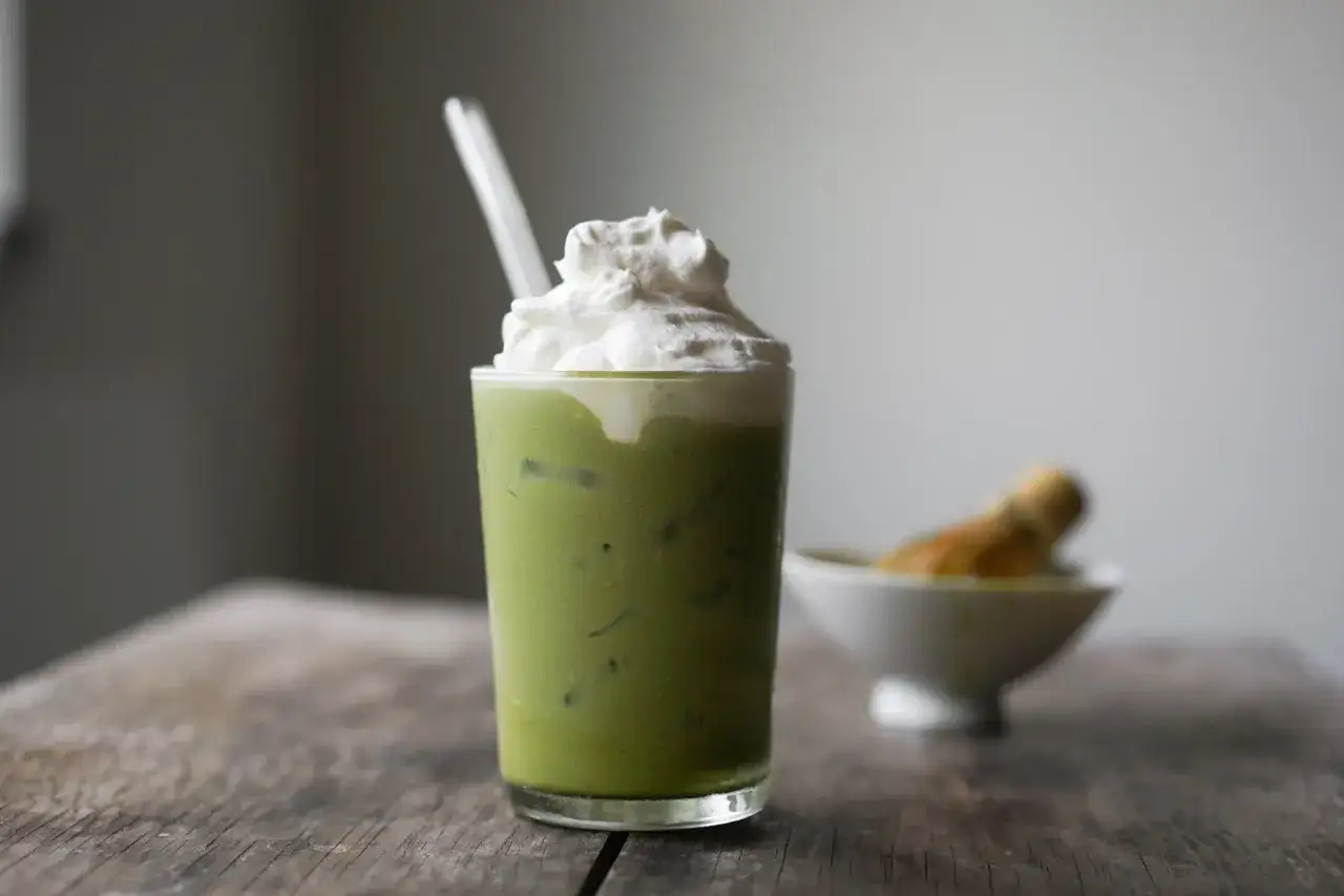 Ein gr&uuml;nes Dalgona Matcha Getr&auml;nk mit Sahnehaube und Eisw&uuml;rfeln, daneben eine Schale mit einem Matcha-Besen.