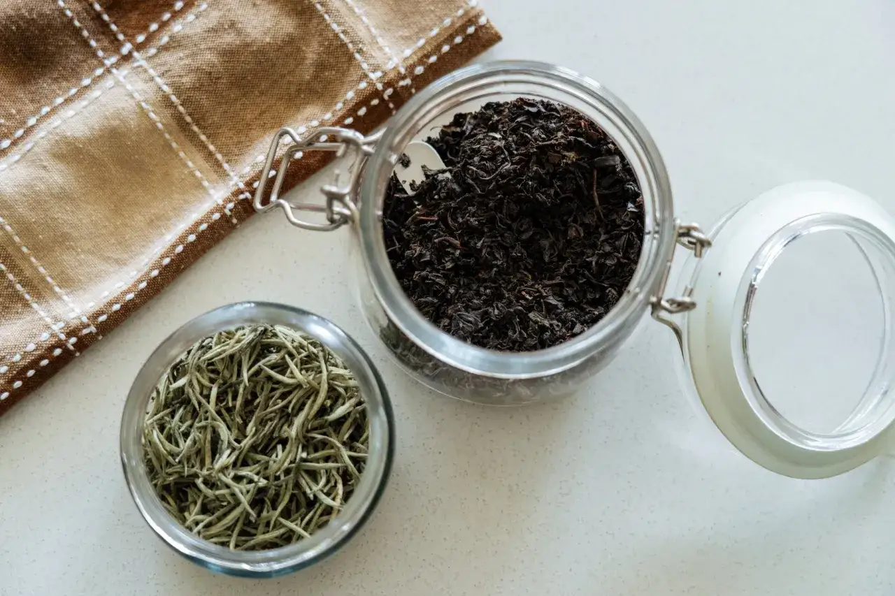 Zwei Gl&auml;ser mit losem Tee: dunkler schwarzer Tee, reich an Koffein, und heller gr&uuml;ner Tee. Daneben ein kariertes Tuch.