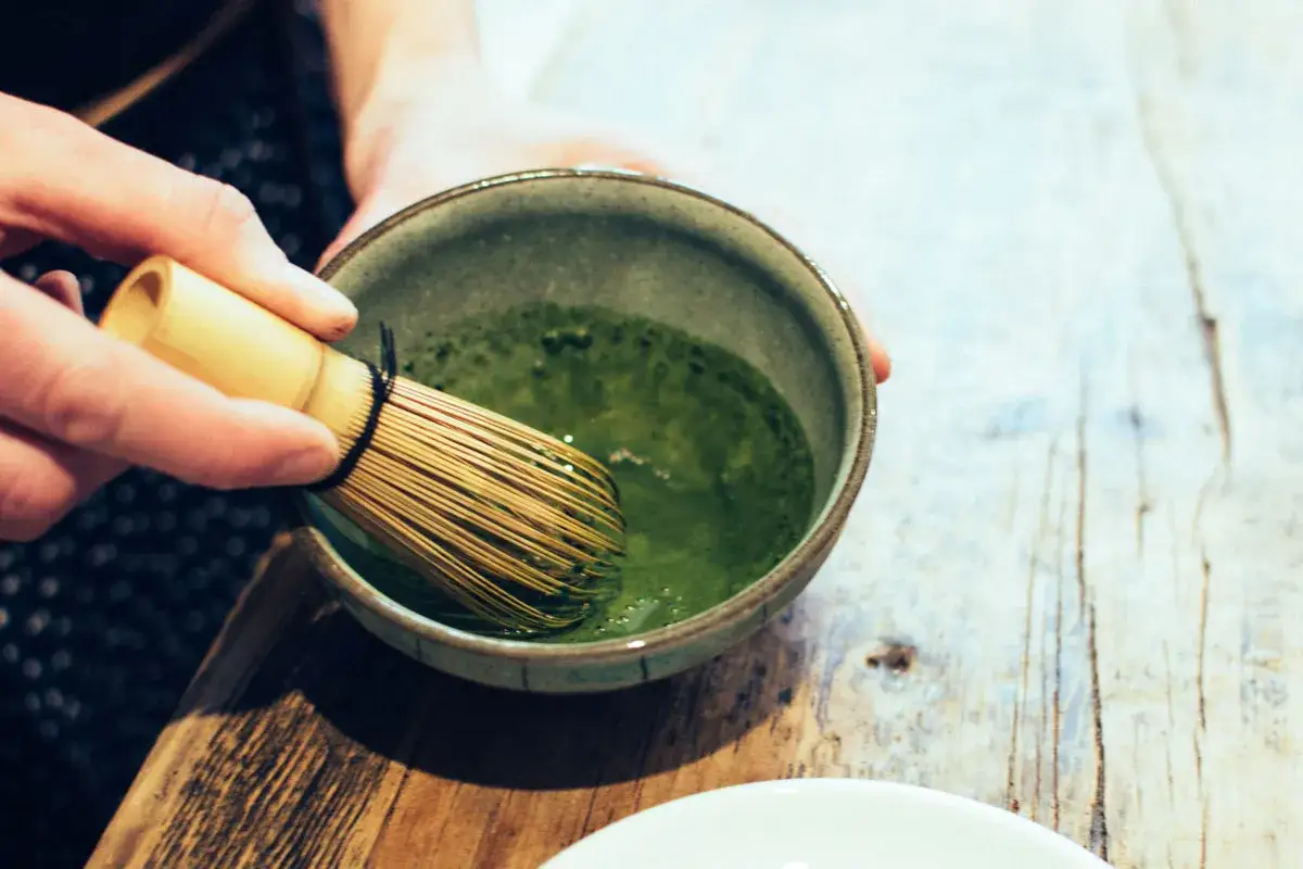 Eine Hand hält einen Bambusbesen, um Matcha-Pulver in einer Schale zu verrühren. Die leuchtend grüne Farbe und die feine Textur des Tees unterstreichen die besonderen Matcha-Eigenschaften.
