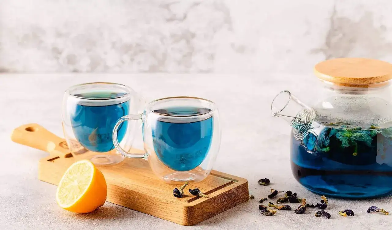 Zwei Tassen leuchtend blauer Tee, eine Teekanne und eine Zitronenspalte auf einem Holzbrett. Informiere dich über blauer tee nebenwirkungen.