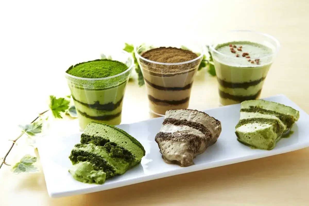 Drei Schichten-Desserts, darunter ein gr&uuml;nes Matcha-Dessert, und zwei Kuchenst&uuml;cke auf einer wei&szlig;en Platte.