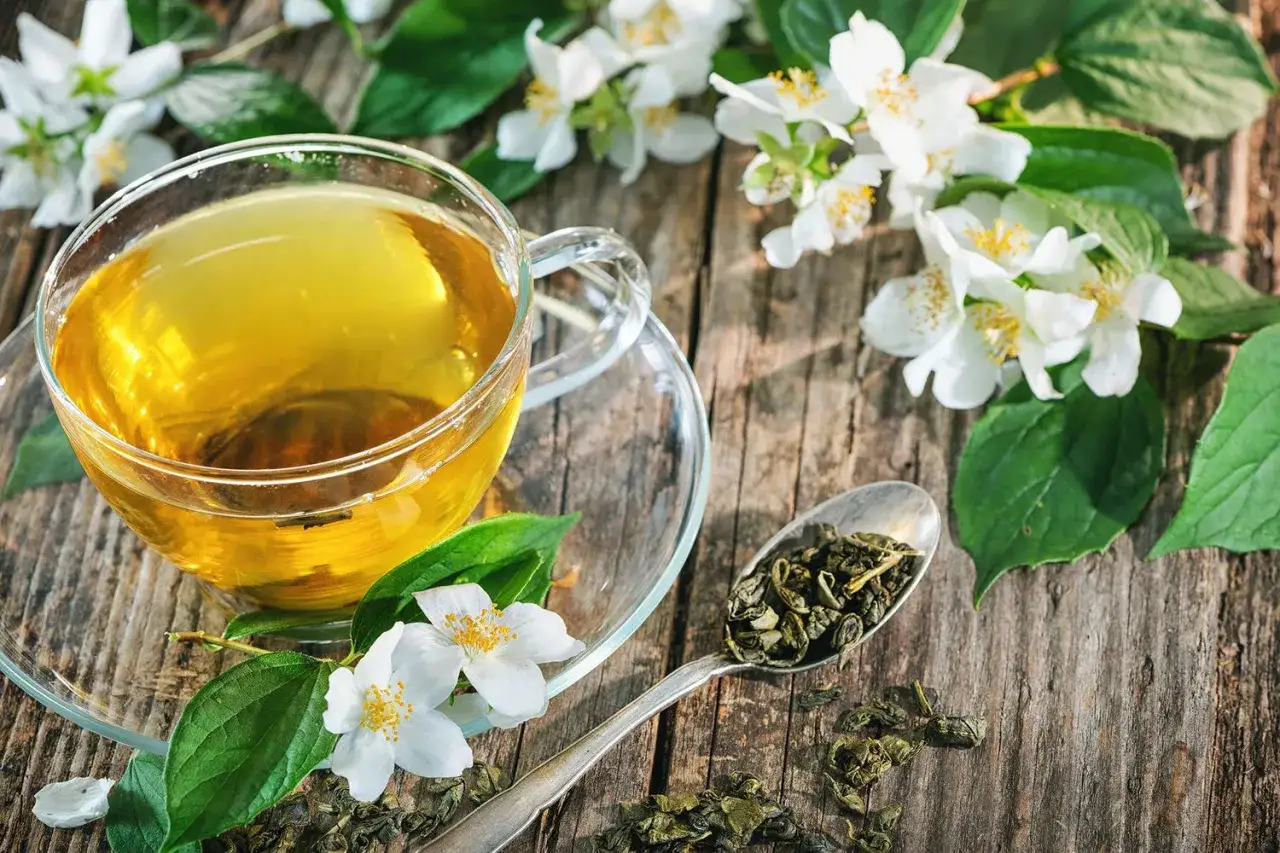Eine Tasse goldener Jasmintee neben frischen Blüten und einem Löffel Teeblätter. Jasmintee gesund und erfrischend.
