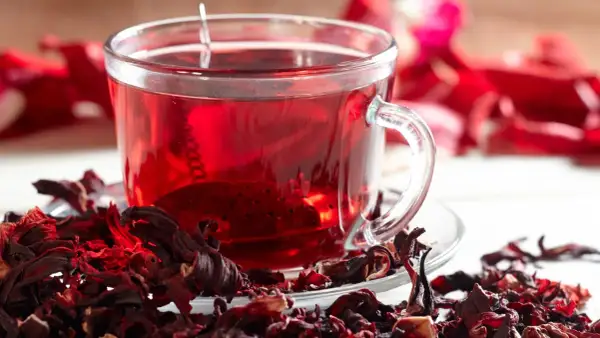 Ein Glas leuchtend roter Hibiskustee, umgeben von getrockneten Hibiskusblüten. Ist Hibiskustee gesund? Ja, er ist voller Antioxidantien.