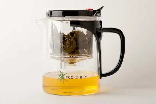 Glas-Teekanne mit schwarzem Deckel und Griff, gefüllt mit goldenem Tee und grünen Teeblättern. Perfekt, um Tee zu servieren.