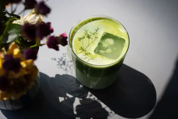 Ein Glas grüner Matcha mit Eiswürfel und einer Pflanze darauf. Neben dem Glas steht ein Strauß getrockneter Blumen. Mögliche matcha nebenwirkungen sind hier nicht ersichtlich.