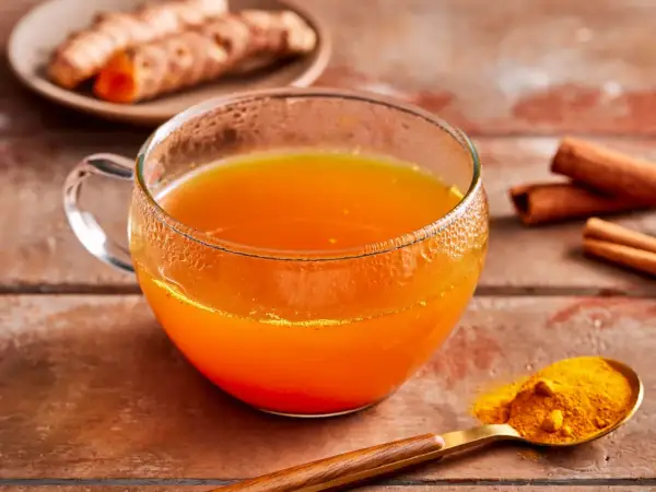 Ein Glas goldener Kurkuma Tee, daneben frische Kurkumawurzeln, Zimtstangen und ein Löffel Kurkumapulver. So einfach lässt sich Kurkuma Tee zubereiten.